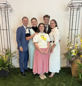 The image shows the Pastor, Joshua Carney, his wife, Rebecca, and their three children, Judah, Chiaryn, and Pemberly standing on a faux grass rug with decorative wire columns and potted flowers on either side.